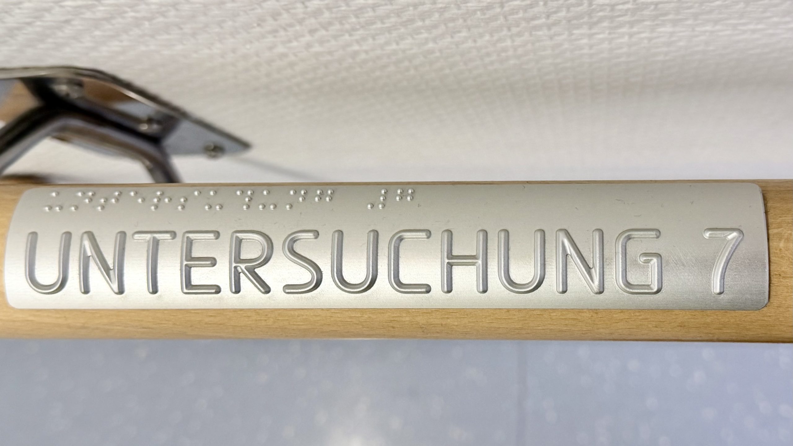 Raumnummern für alle. Handlauf mit einer tastbar erhabenen Beschriftung "Untersuchung 7" in Druckbuchstaben und Braille.